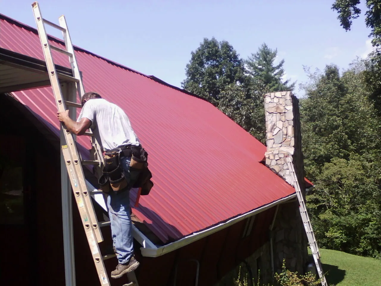 Roofer on a ladder beside a red metal roof and stone chimney — Soffit & Fascia Repair by Roofing Brothers in Williams Bay